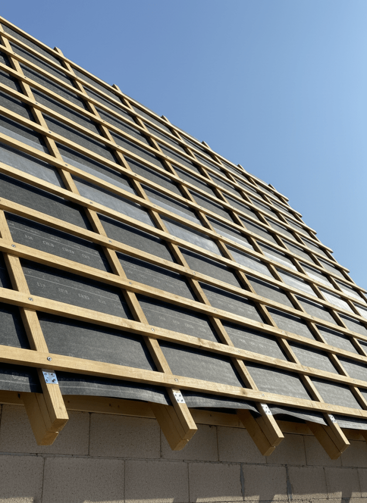 A detailed view of a pitched roof structure in progress, showing precisely arranged light pine rafters and battens forming a geometric pattern over a solid, insulated underlay membrane in dark gray. High-quality vapor barrier sheets are neatly fixed, edges perfectly straight, with metallic fixing plates and screws aligned with engineer-like regularity. The structure sits above a partially visible neutral-toned masonry wall. The scene is bathed in bright but slightly diffused midday sunlight, emphasizing the wood grain and material contrasts without harsh glare. Captured from a low-angle perspective looking up along the roof slope to highlight stability and strength, with a clear blue sky providing a clean background. The mood is technical and trustworthy, with a modern, documentary photographic realism ideal for a professional roofing company.